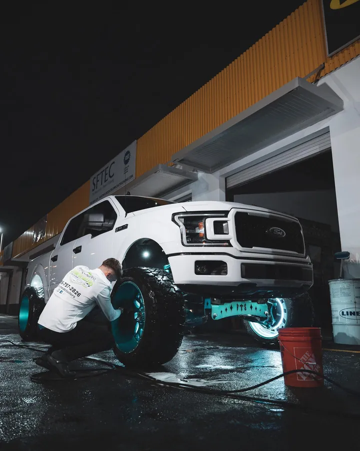 Divine Detailers technician hand-washing the custom wheel of a lifted white Ford F-150 at night