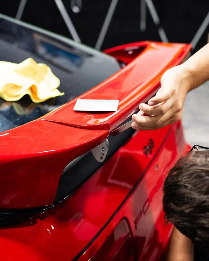 Hands lifting paint protection film along the edge of a red Porsche 968 rear spoiler
