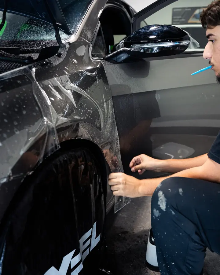 Technician smoothing clear paint protection film onto the fender of a gray car