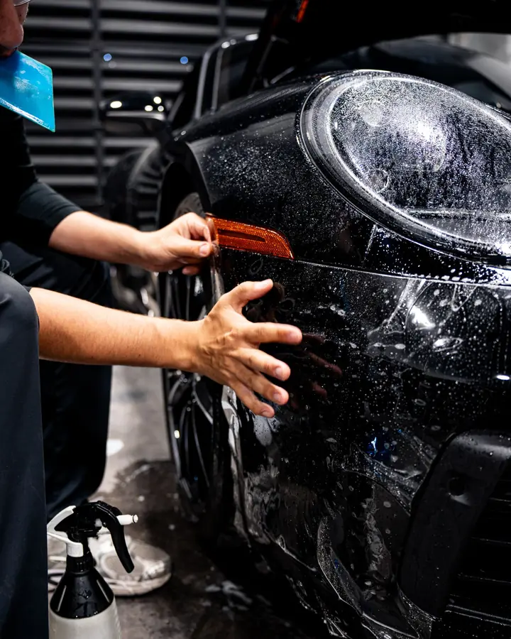 Hands smoothing clear paint protection film around the corner marker light of a black Porsche