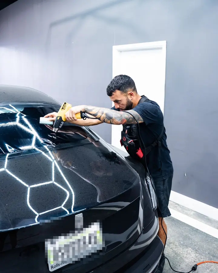 Technician using a heat gun and squeegee to install window tint on the rear glass of a black luxury car