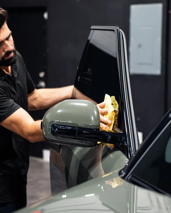 Technician cleaning around the side mirror and tinted window of an olive green SUV