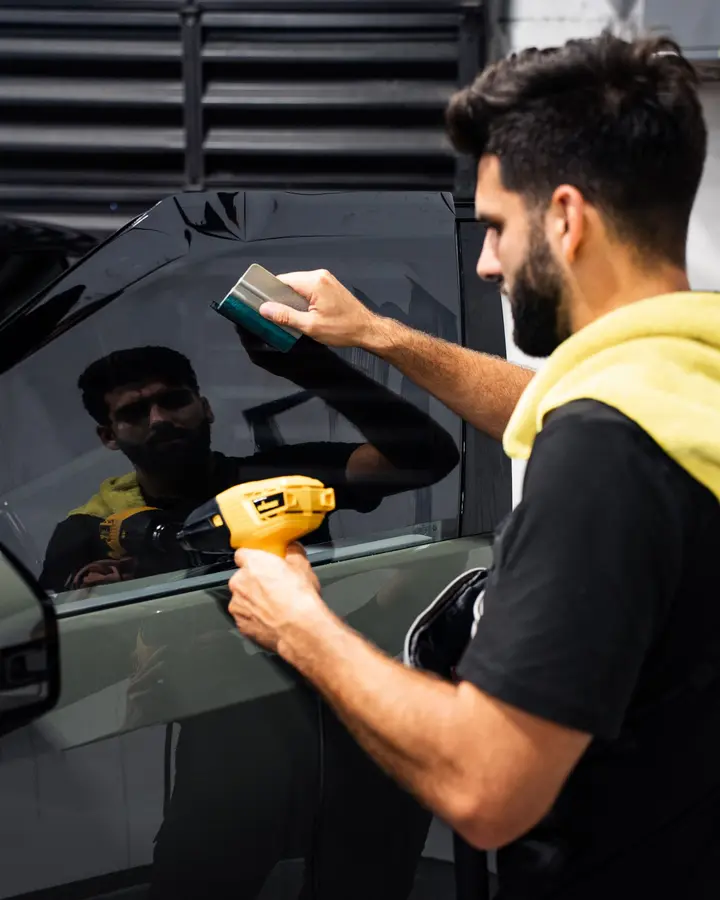 Technician using a squeegee and heat gun to install window tint on an olive green truck